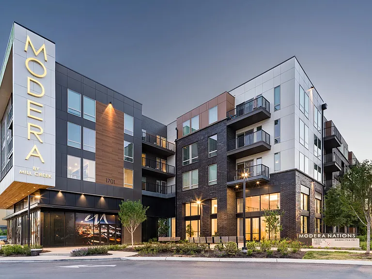 Modera Nations features bold architecture, mixed materials, and illuminated signage anchoring the neighborhood streetscape at dusk.