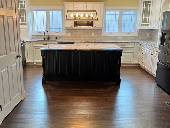 View from great room to kitchen with large island with lots of storage below and new lighting fixture for plenty of lighting in the kitchen. New custom made cabinets and light white/grey brick backsplash. New large windows and door leading to basement.