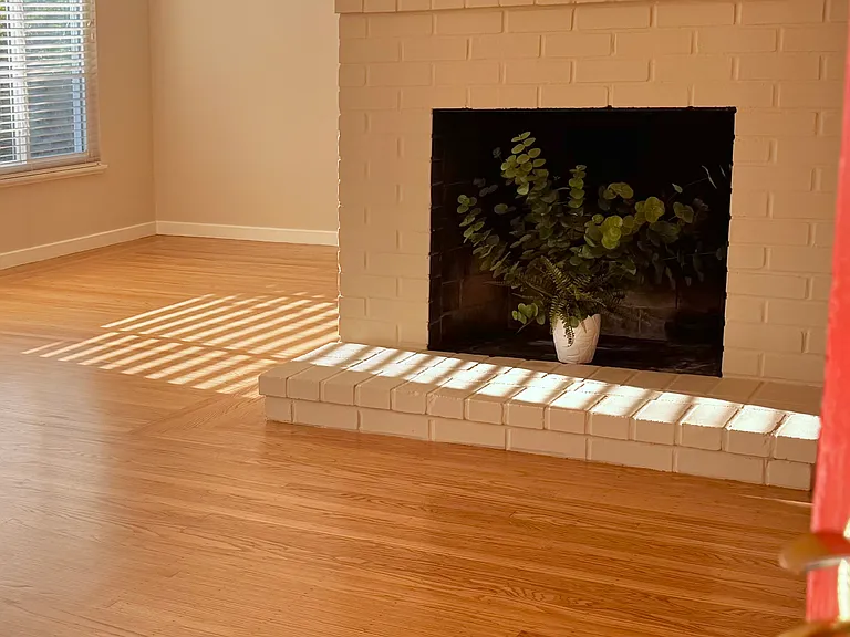 Step inside to a light-filled living area featuring warm, newly refinished hardwood floors, fresh paint, clean mid-century lines, and a brick fireplace as the central focal point.