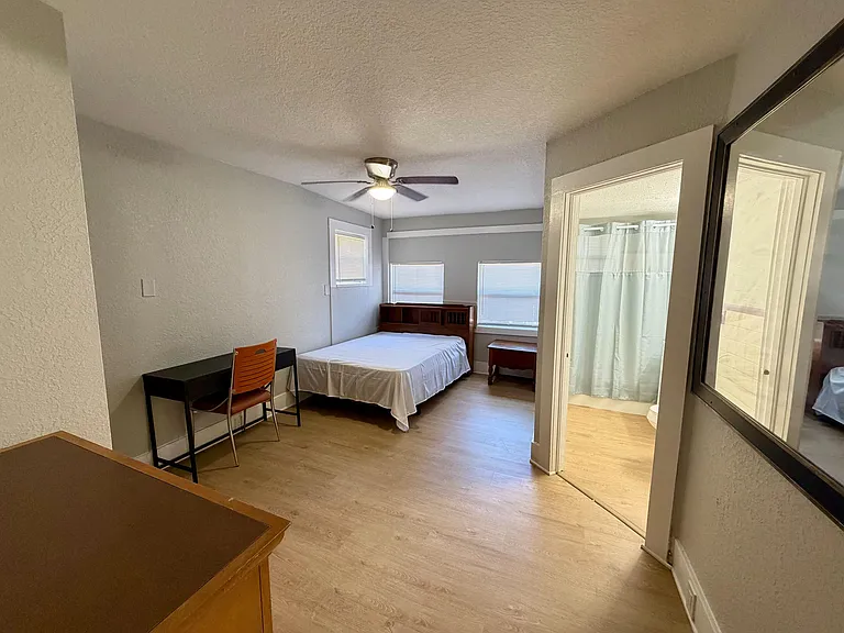 Furnished bedroom with a full bed, desk workspace, ceiling fan, and two windows for natural light; bath access nearby.