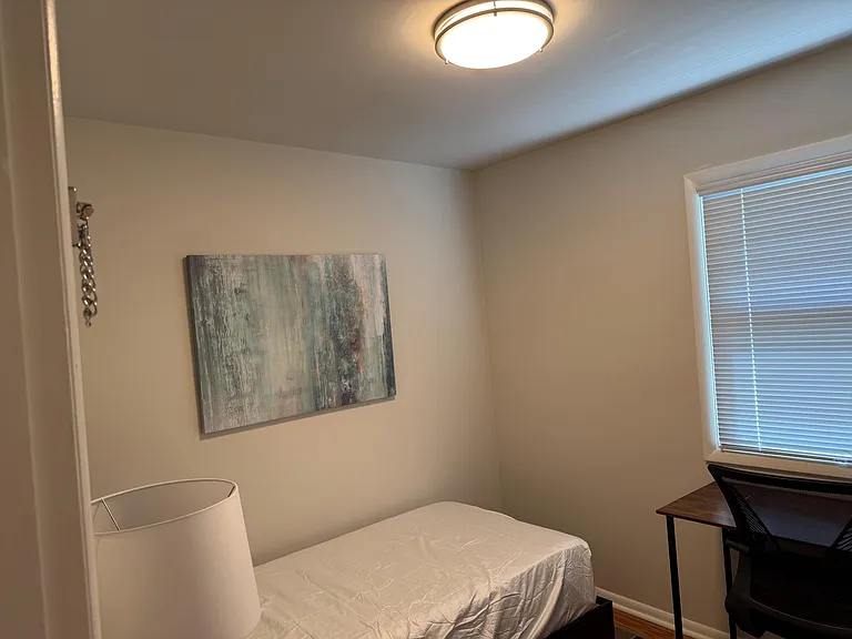 Private bedroom with a twin bed, overhead lighting, and a window with blinds, plus a compact desk area for work or study.