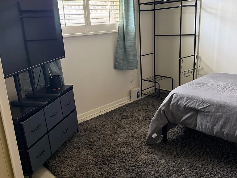 Bedroom with a window and plantation shutters, space for a bed, and convenient storage with dresser and open shelving.