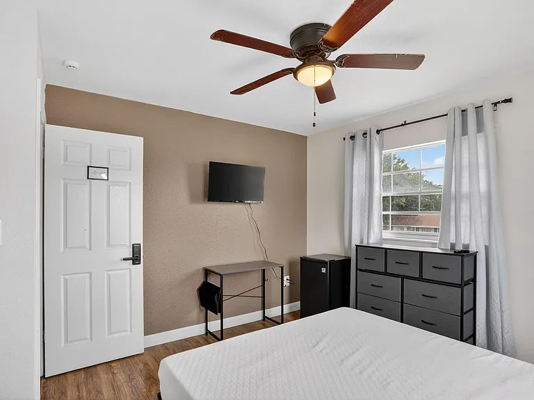 Bedroom includes a ceiling fan, window with curtains, dresser storage, and a mini fridge with space for a small desk.