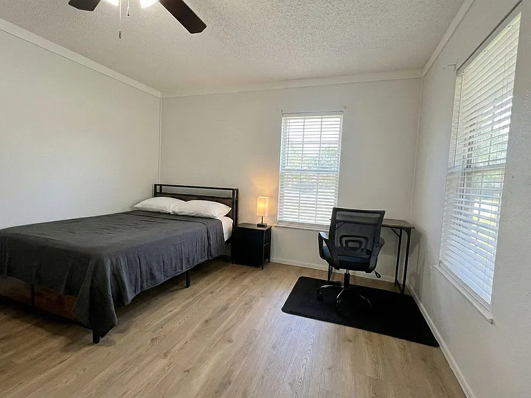 Spacious bedroom with a queen bed, wide-plank flooring, two windows with blinds, and a dedicated desk workspace.