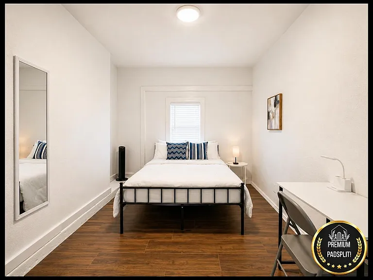 Furnished bedroom with a full bed, work desk, and floor mirror, plus wood-style flooring and a window with blinds.