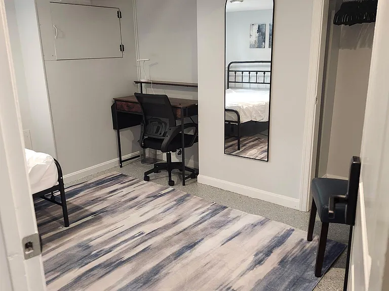 Private bedroom with a work desk, full-length mirror, and open closet storage, plus space for a bed and seating.