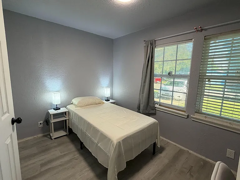 Private bedroom with twin bed, nightstands, and large windows with blinds and curtains for natural light and privacy.
