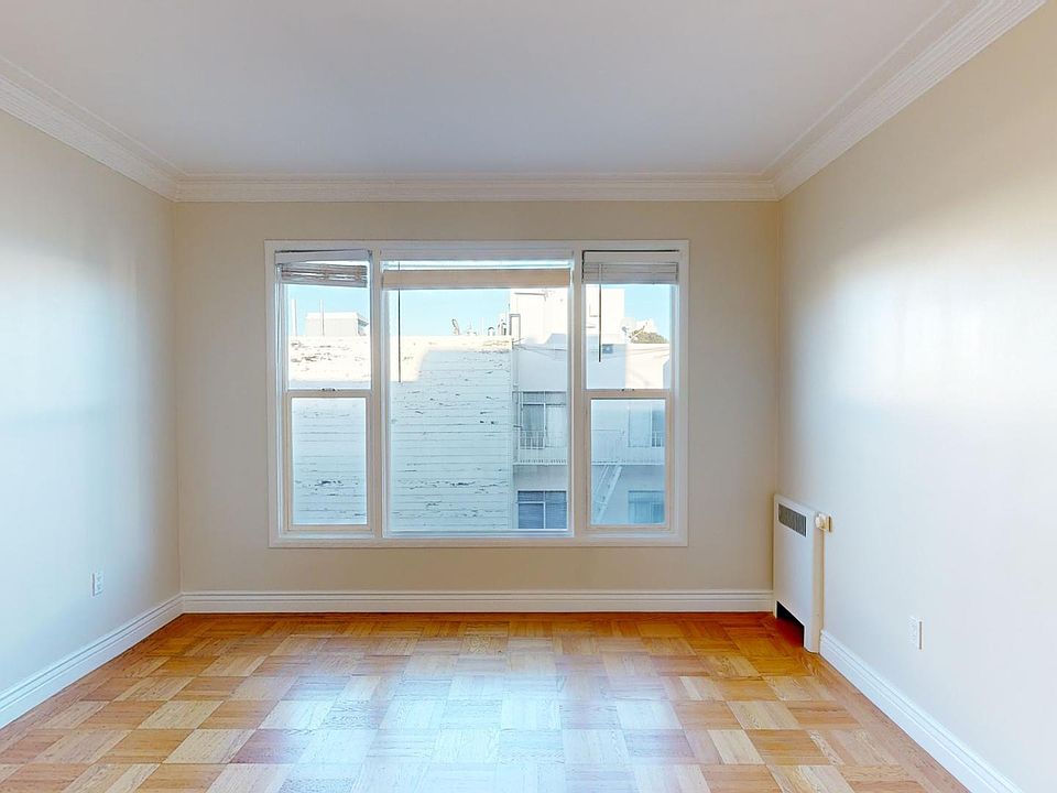 Bright, empty room with hardwood flooring and large windows, offering a view of neighboring buildings against a clear sky.