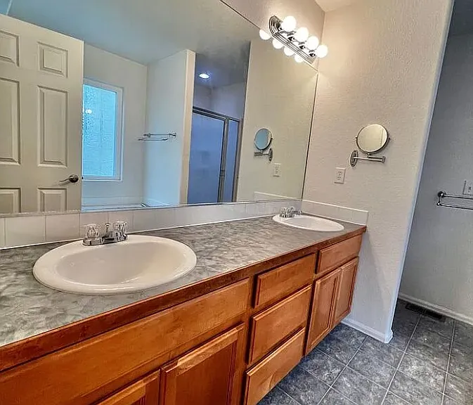 Dual sinks and cabinets with a large mirror along the northern master bathroom wall. The toilet is located in an alcove on the other side of the wall seen just past the sink area. To the right (not visible) are the large shower and garden tub.