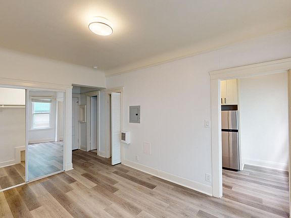 Room with light wood flooring, mirrored closet, and visible kitchen area