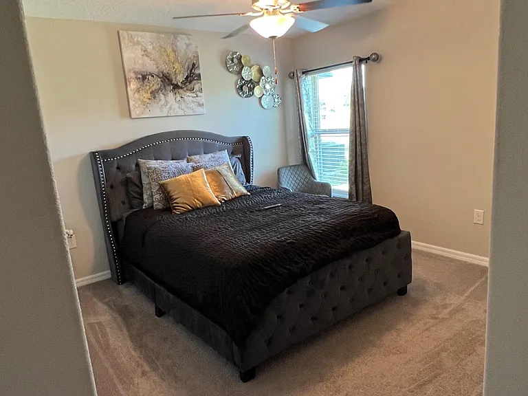 Carpeted bedroom featuring a tufted bed, ceiling fan with light, and a window with curtains for natural daylight.
