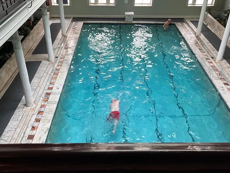 Indoor lap pool area with tiled deck, column-lined perimeter, and upper-level balcony overlooking the water.