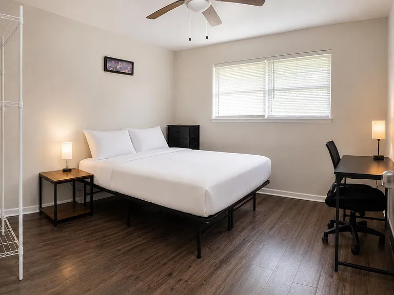 Furnished bedroom with a queen bed, desk workspace, large window with blinds, and open shelving for storage.