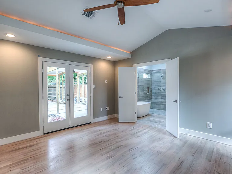 Vaulted master bedroom with double French doors to the backyard, cove lighting, and view into a spa-style master bath with freestanding tub
