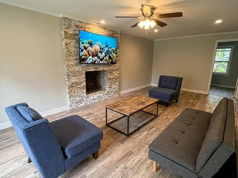 Spacious living room with durable plank flooring, recessed lighting, and a ceiling fan, anchored by a stone fireplace wall.
