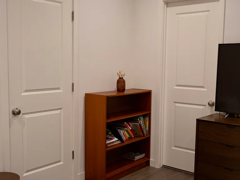 Bedroom 2: Captures the wall layout featuring the main room door and the ensuite bathroom door, with a corner bookshelf perfectly positioned in between.