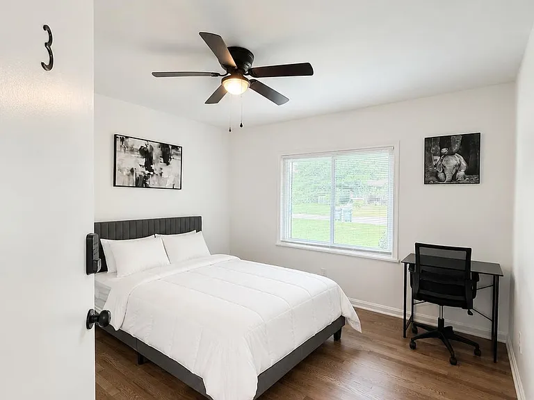 Furnished bedroom with hardwood floors, ceiling fan, large window, and a dedicated desk workspace.