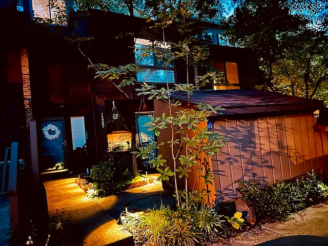 As evening falls, the front entrance glows softly, framed by the gentle silhouette of a private azalea garden. Subtle outdoor lighting highlights the blooms and greenery, creating a tranquil, welcoming atmosphere.