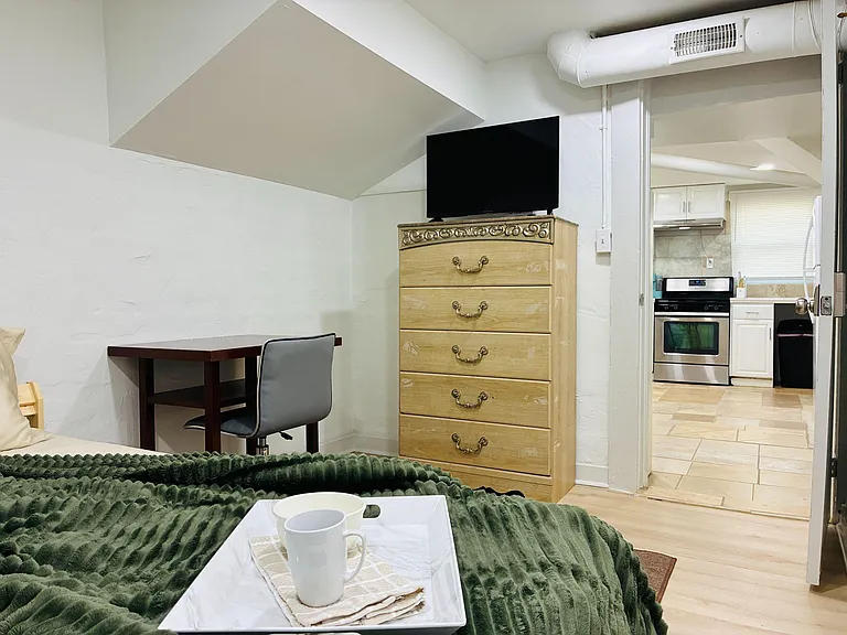 Furnished bedroom with a work desk, dresser with TV, and easy access to the adjacent kitchen for added convenience.