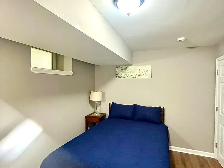 Private bedroom with a full bed, nightstand and lamp, plus hardwood-style flooring and overhead lighting.