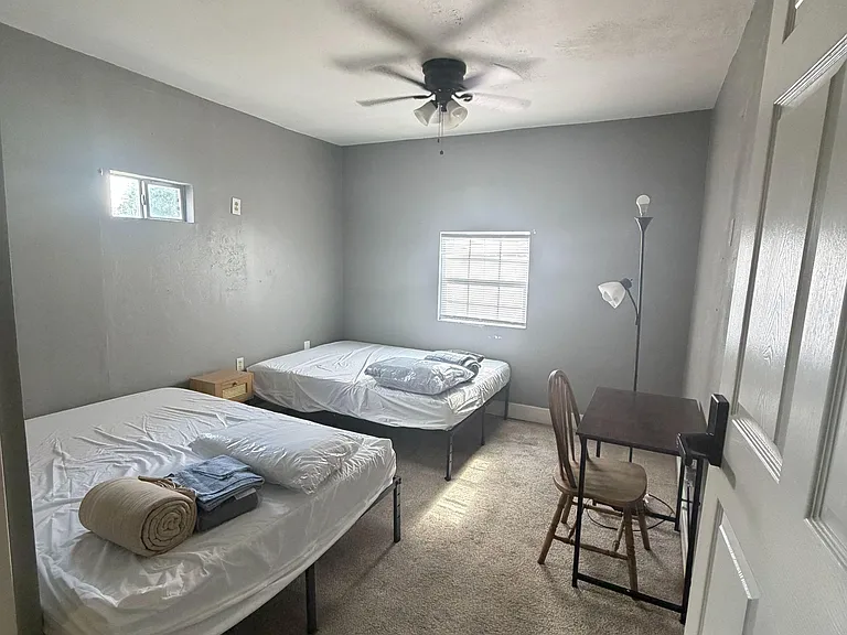 Shared bedroom with two twin beds, carpeted floors, ceiling fan, and a desk setup with natural light from two windows.