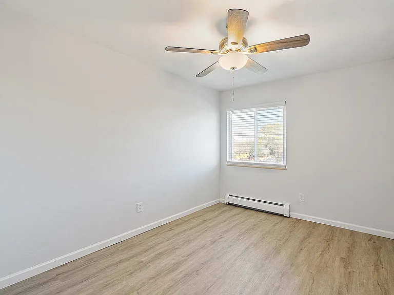 Bright and airy room with modern ceiling fan and large window, featuring sleek laminate flooring and fresh white walls.