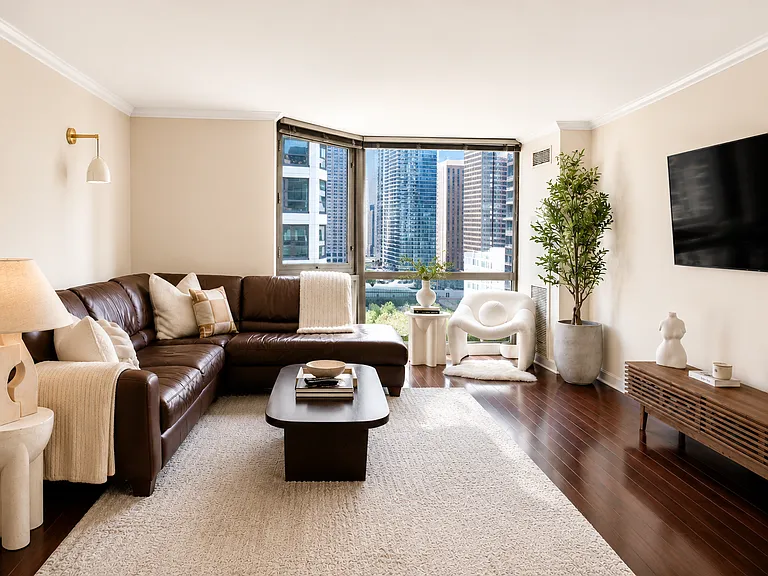Sun-drenched living area with rich dark hardwood floors and bay windows showcasing stunning skyline views. The open layout provides generous space for a large sofa and entertainment center, perfect for relaxing or hosting guests.