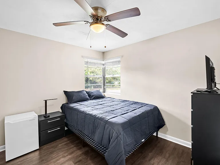 Furnished bedroom with a queen bed, corner windows, ceiling fan, and space for a dresser or small work setup.