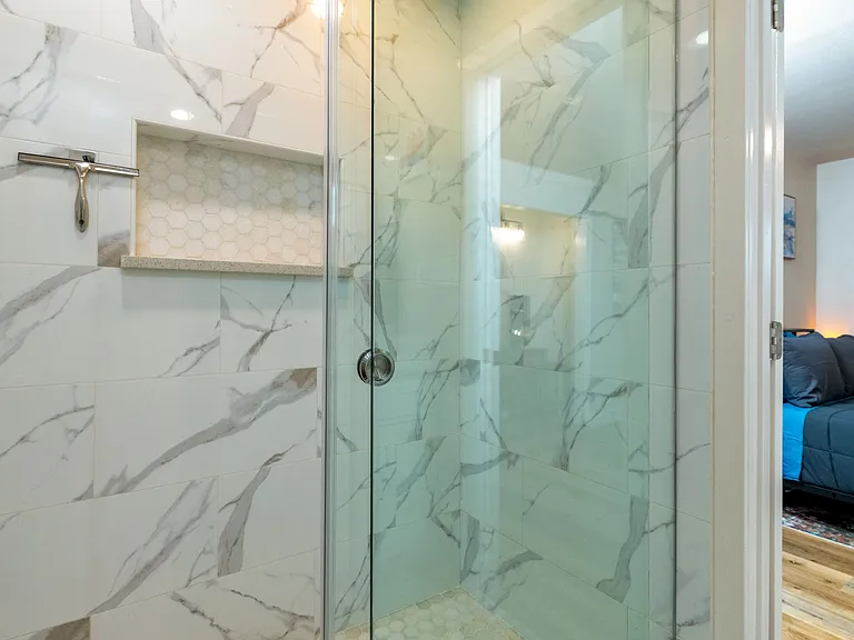 Relax in a modern, spa-style bathroom with a marble-tiled walk-in shower, sleek black fixtures, bright vanity lighting, and clean finishes. Directly connected to the primary bedroom for privacy and convenience.