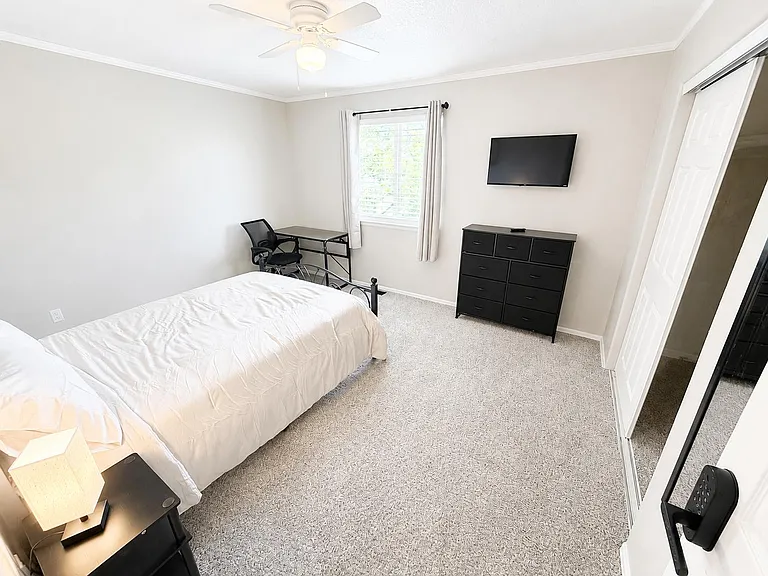 Furnished bedroom with a full bed, desk, dresser, and wall-mounted TV, plus a window with blinds and ceiling fan.