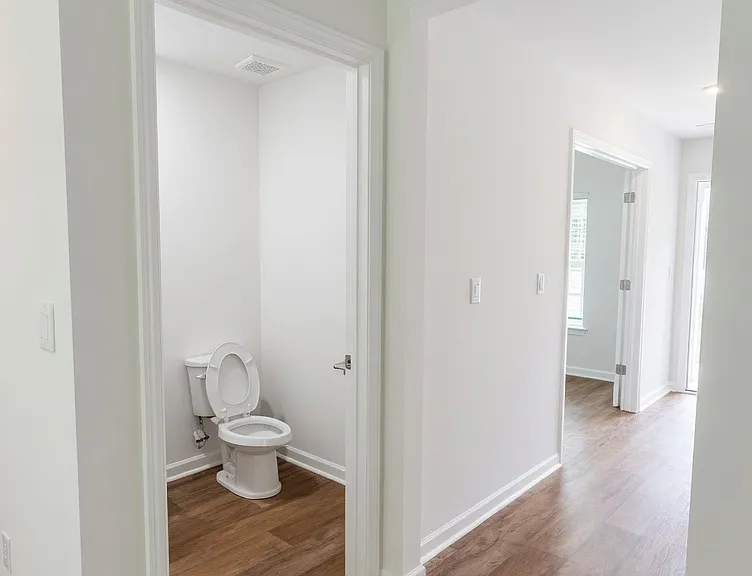 Wide hallway with wood-look flooring and crisp white walls and trim. Powder room (half-bath) is immediately to the left with a white toilet and simple finishes.