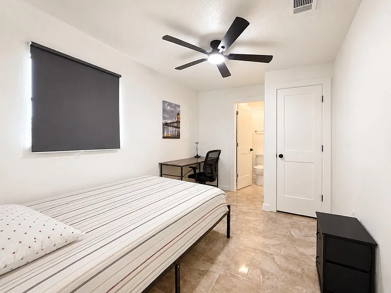 Private bedroom with a large window, ceiling fan, blackout shade, tile flooring, furnished with a large closet and work desk.