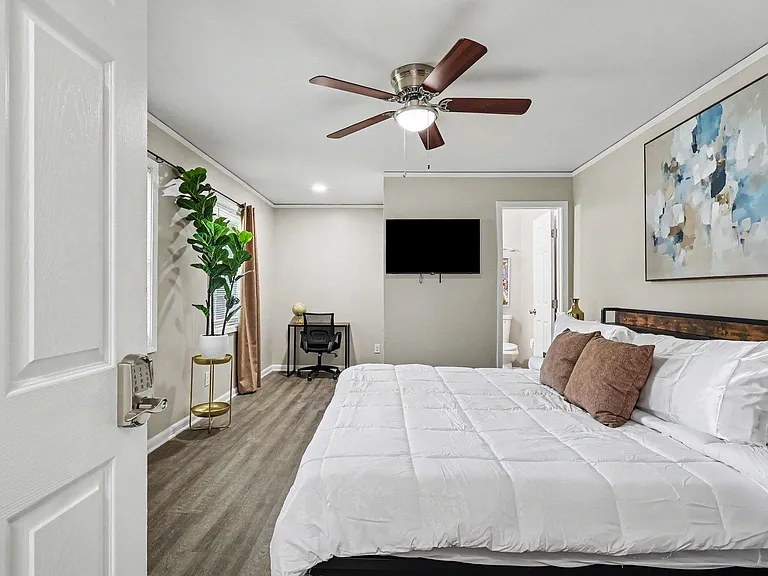 Spacious furnished bedroom with a ceiling fan, wall-mounted TV, and work desk; wood-look flooring and an ensuite bath entry.