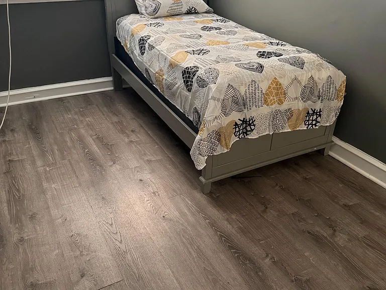 Private bedroom with a twin bed, durable wood-look flooring, and a window with blinds plus in-room A/C for comfort.