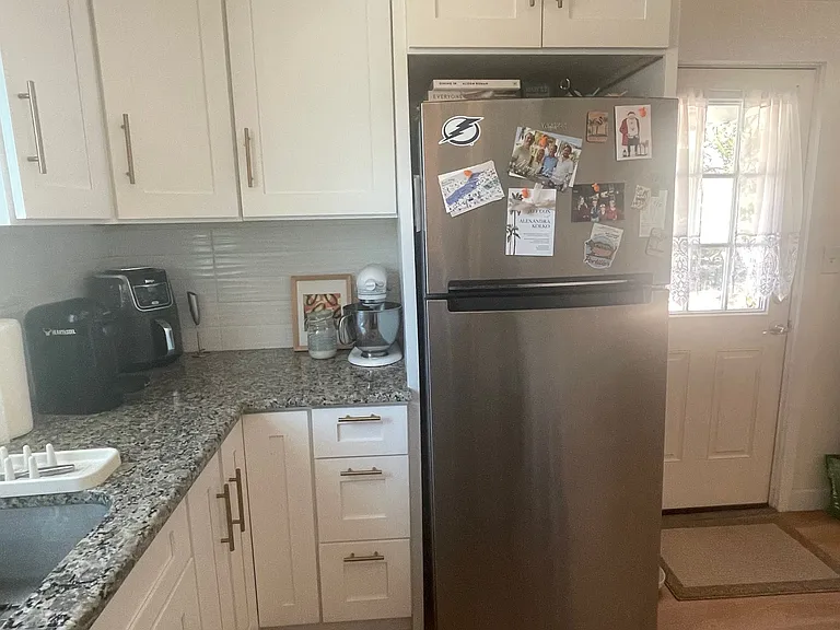 Kitchen area has a stainless steel refrigerator, and white shaker cabinets with ceiling moldings. All fixtures are brass/gold tone - kitchen faucet, cabinet pulls, and light fixture. Ceiling fan in LR also has gold accent.