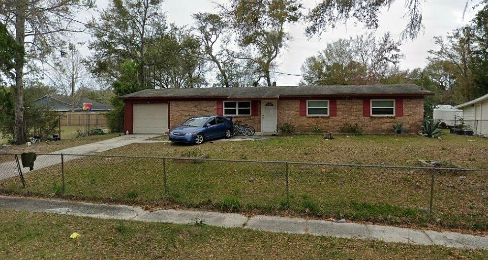 Brick home with large fenced yard, attached garage, long driveway, and shade from mature trees. Red-trimmed windows and door enhance the inviting, open feel of this single-story residence.