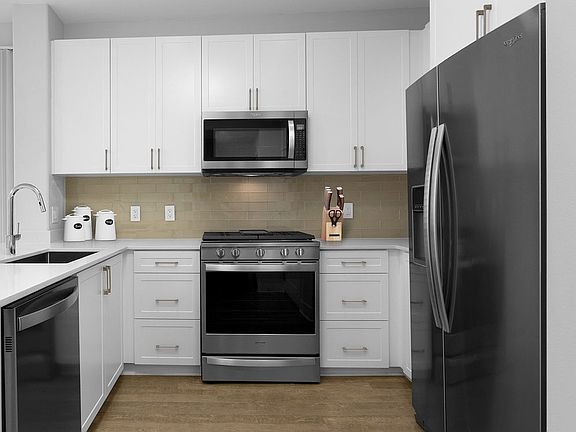 Renovated Package I kitchen with stainless steel appliances, white speckled quartz countertops, white cabinetry, grey tile backsplash, and hard surface flooring (Representative Imagery)