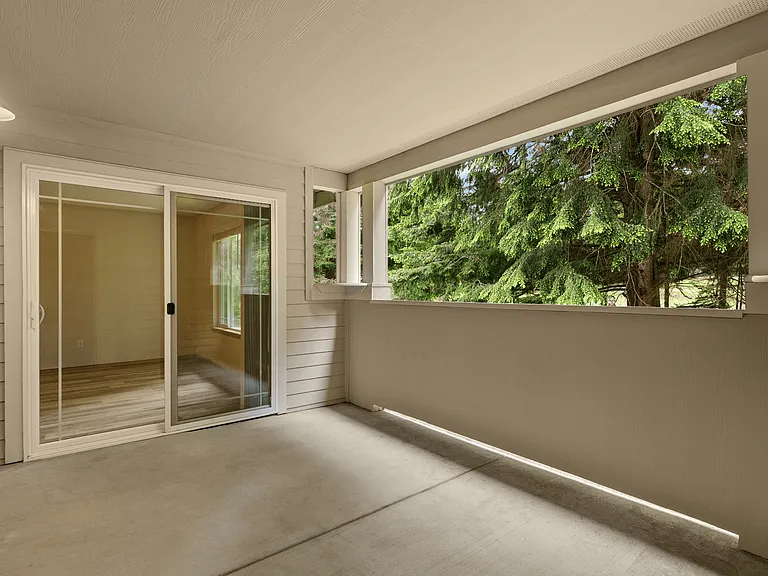 Covered patio with concrete flooring, beige siding, and a large open window space overlooking green pine trees, accessed by a sliding glass door. Covered patio with concrete flooring, beige siding, and a large open window space overlooking green pine tree