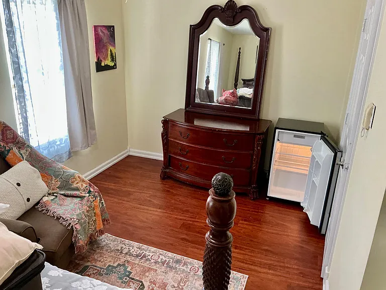 Bedroom with wood-look flooring, a dresser with mirror, window with curtains, and an in-room mini fridge for added convenienc