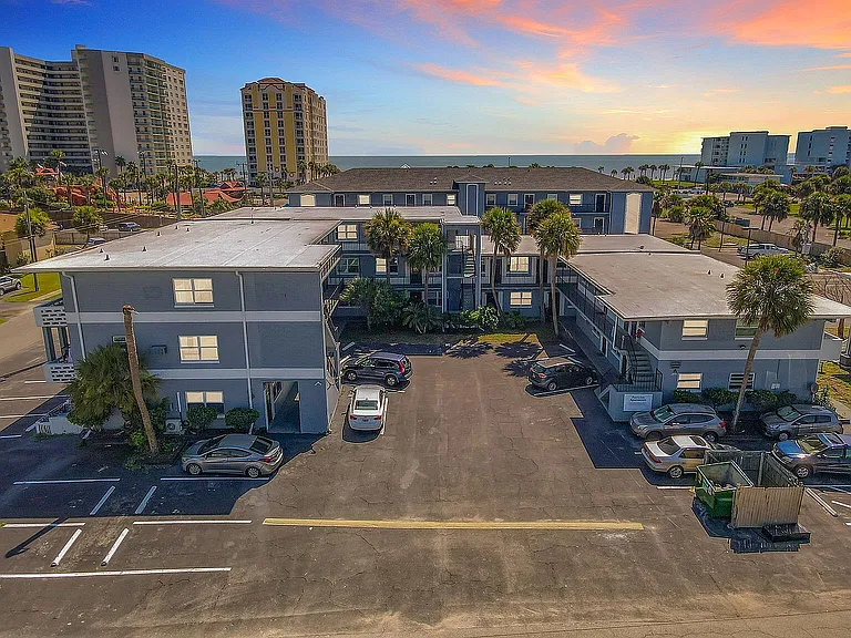 Modern coastal apartment complex with ample parking and vibrant sunset views, surrounded by lush palm trees and sky-touching beachfront towers.