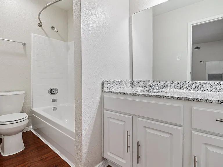 Bright and modern bathroom with sleek speckled countertop, white cabinetry, and polished wood-style flooring, featuring a bathtub and shower combo. Bright and modern bathroom with sleek speckled countertop, white cabinetry, and polished wood-style floorin