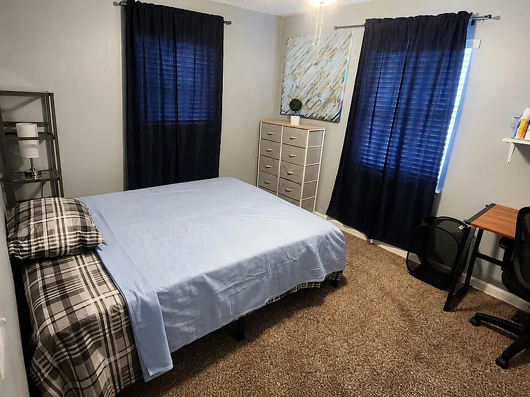 Furnished bedroom with a Queen bed, dresser, and dedicated desk area, plus two windows with blinds and curtains for privacy.