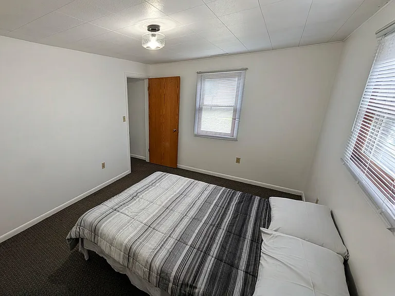 Private bedroom with a full-size bed, carpeted floors, and two windows with blinds for natural light and privacy.