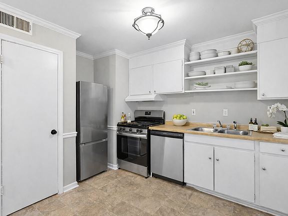 This bright, thoughtfully designed kitchen features crisp white cabinetry, open shelving, warm countertops, stainless steel appliances, and a clean, airy layout that feels functional, welcoming, and perfect for everyday living and entertaining.