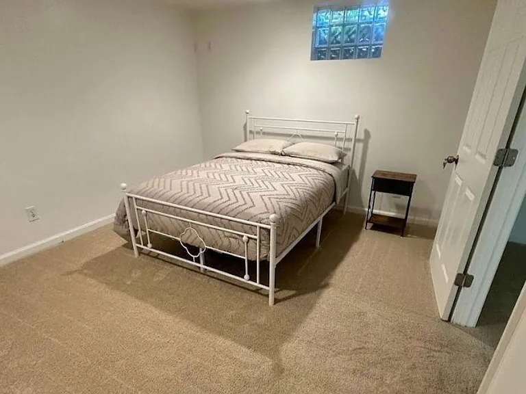 Furnished bedroom with carpeted floors, a full bed, and a nightstand, plus a glass-block window for natural light.