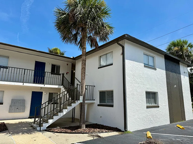 Charming white apartment building with modern railings, surrounded by lush palm trees under a bright blue sky.