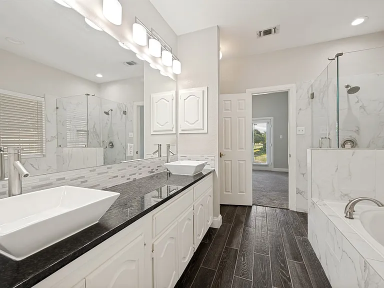 Spacious bathroom featuring dark wood-finish flooring, a double vanity with vessel sinks and dark countertops, a tiled walk-in shower with glass enclosure, and a built-in soaking tub with tile surround