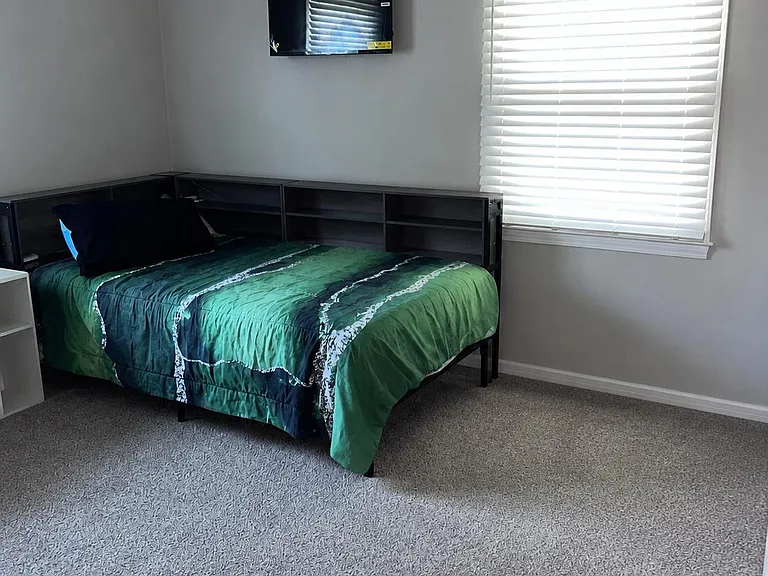 Furnished bedroom with a twin bed, wall-mounted TV, and a large window with blinds for natural light and privacy.