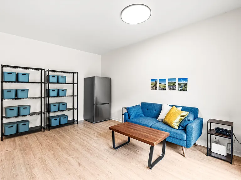 Shared lounge with a sofa and coffee table, plus open shelving for groceries and a full-size refrigerator for added storage.