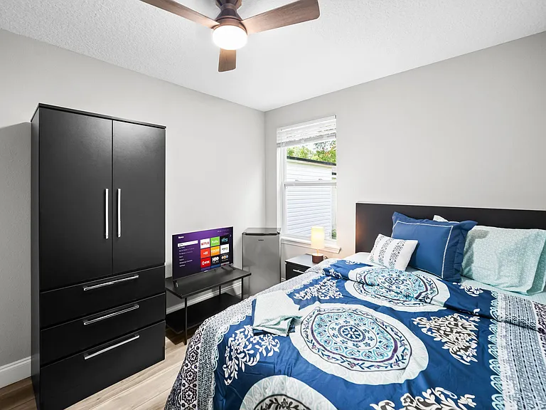 Furnished bedroom with a full bed, dresser storage, TV stand, and a mini fridge, plus a window and ceiling fan.